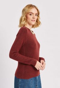 Sarkans adīts džemperis ar apaļu kakla izgriezumu un garām piedurknēm, ar teksturētu apdari un ribotām aprocēm. Modelis valkā dekoratīvu kaklarotu.