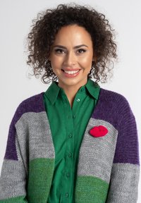 Sweter z dzianiny w paski w odcieniach szarości, zieleni i fioletu, w połączeniu z zieloną koszulą. Czerwona pleciona broszka w kształcie ust dodaje detalu.