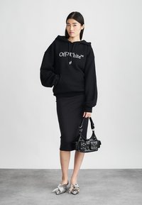 Črna pulover s kapuco z belim logotipom "Off-White™", v kombinaciji s prilegajočo črno krilo, čevlji z srebrnimi detajli in črno torbico s studenci.