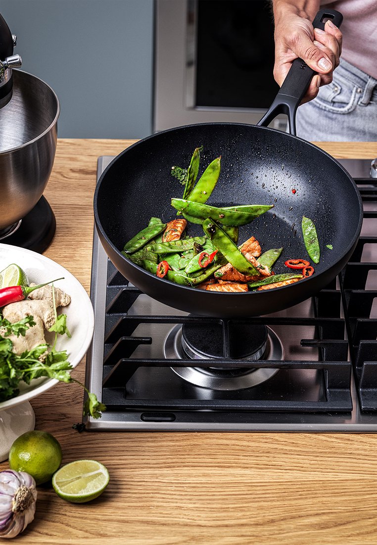 Schwarzer antihaftbeschichteter Wok auf einem Gasherd mit angebratenen Zuckerschoten, roten Chilischeiben und Lachsstücken. Holztresen mit Kräutern und Limetten in der Nähe.