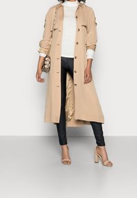 Beige langer Trenchcoat mit Knöpfen, über einem weißen Rollkragenpullover getragen, kombiniert mit schwarzen Hosen und beige High-Heels. Kleine Handtasche mit Muster.