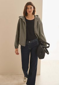 Světle zelená bunda na zip s kapucí, černé tričko a tmavě modré široké džíny. Drží černou nafukovací kabelku. Na nohou má ploché tenisky.