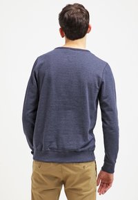 Muž stojící zády, oblečený v dlouhém modro-navy svetru a béžových kalhotách, zasazený na pozadí prosté bílé stěny.