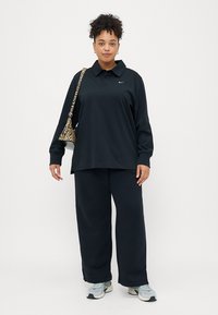 Crna majica s dugim rukavima i ovratnikom, s Nike logom, u kombinaciji s širokim crnim hlačama i torbom s uzorkom. Model nosi tenisice.