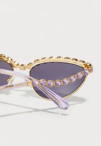 Occhiali da sole a occhio di gatto con lenti grigie scure, montatura dorata, bordo rialzato e accenti di strass lavanda sulle tempie e sulle aste.