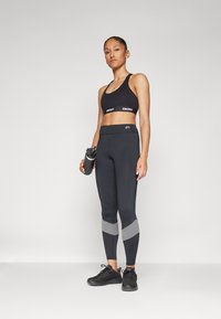 Soutien desportivo preto com a marca DKNY, leggings altas pretas com detalhes cinzentos e sapatilhas atléticas pretas. Garrafa de água na mão.