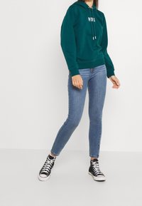 Turkusowy bawełniany hoodie z kieszenią z przodu, sznurkami i detalem logo. Połączony z jasnoniebieskimi obcisłymi dżinsami i czarnymi wysokimi trampkami.