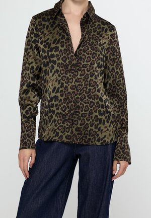 Vrouw draagt een olijfgroene blouse met lange mouwen en luipaardprint en donkerblauwe jeans, handen ontspannen langs het lichaam.