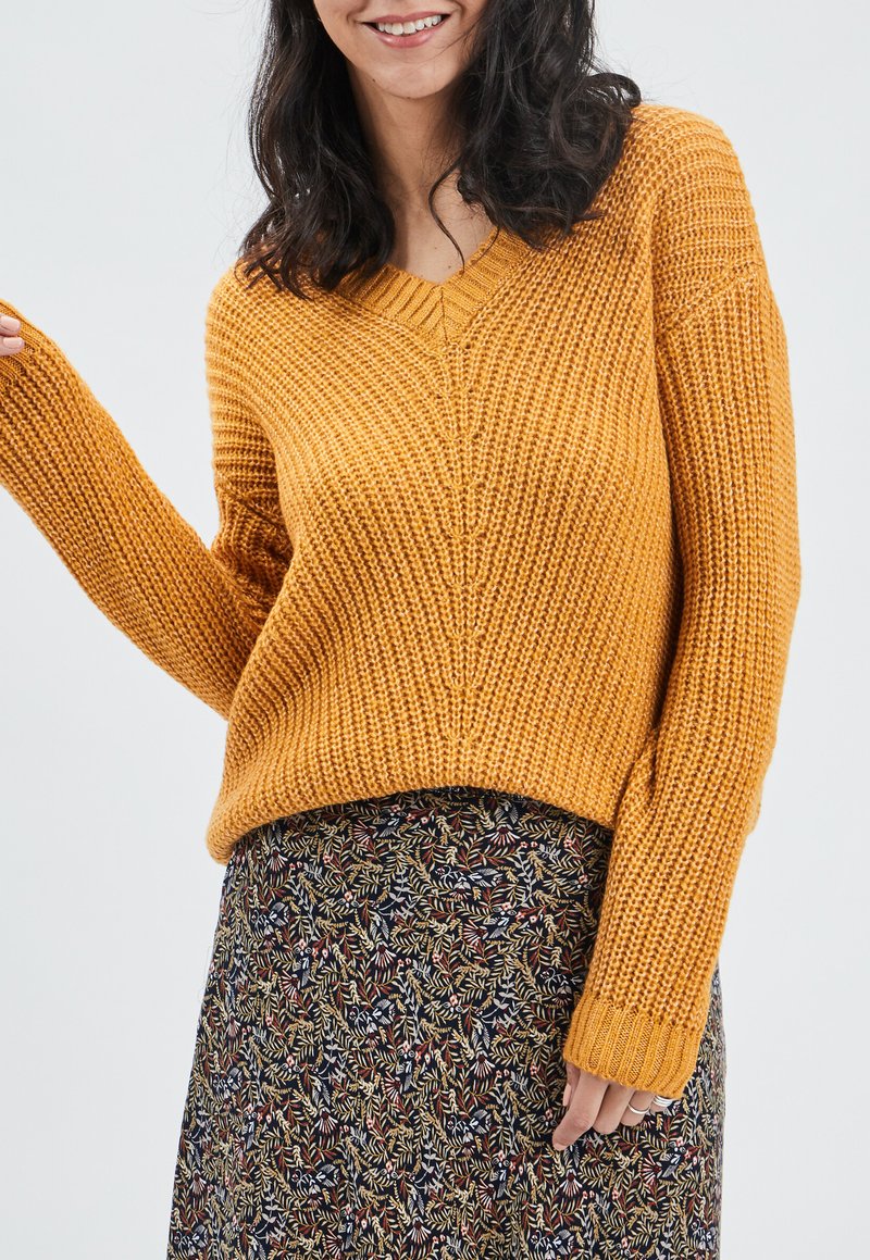 V-Ausschnitt gestrickter Pullover in senfgelb mit lockerer Passform, strukturiertem Stoff und gerippten Bündchen, kombiniert mit einem rock mit Blumendessin.