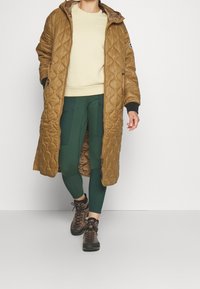 Abrigo largo acolchado marrón con capucha, sudadera beige, leggings verdes y botas de senderismo marrones. Presenta bolsillos y puños contrastantes.