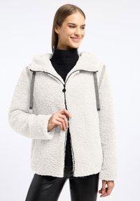 Lichtgrijze fleece jas met een ritssluiting, capuchon en contrasterende grijze banden. Heeft een zachte textuur en een ontspannen pasvorm, gedragen over een zwart bovenstuk.