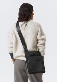 Borsa a tracolla nera con una texture liscia, caratterizzata da una tasca frontale con zip e una tracolla regolabile. Indossata sulla spalla con un maglione chiaro.