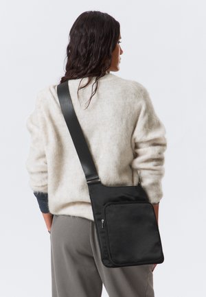 Bolsa transversal preta com uma textura suave, apresentando um bolso com fecho na frente e uma alça ajustável. Usada no ombro, sobre um suéter claro.