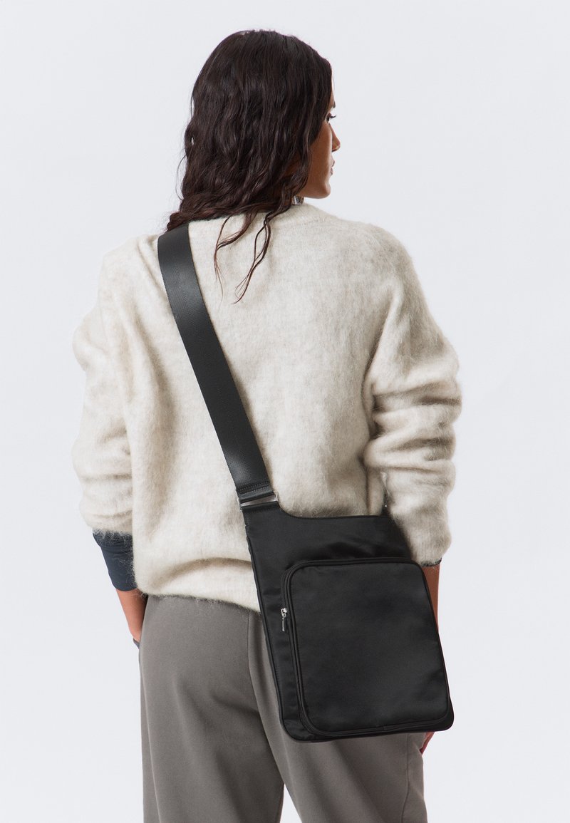 Borsa a tracolla nera con una texture liscia, caratterizzata da una tasca frontale con zip e una tracolla regolabile. Indossata sulla spalla con un maglione chiaro.