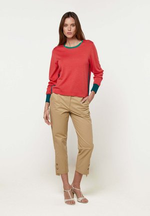 Femme portant un pull rouge avec des bordures vertes, un pantalon beige court avec des détails à boutons, et des sandales blanches à talons hauts, se tenant devant un fond uni.