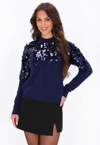 Granatowy sweter z cekinową górą, długimi rękawami i ściągaczami, zestawiony z czarną mini spódnicą z bocznym rozcięciem.