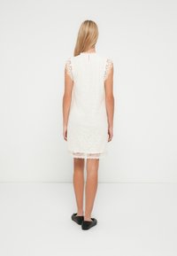 Balta mežģīņu kleita ar īsām, volānu piedurknēm un viļņotu apmali. Aizmugurē ar atslēgas cauruma aizdari un reljefainu ziedu rakstu.