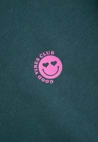 Ciemnozielona bluza z tkaniny z dużą różową grafiką uśmiechniętej buźki z sercowymi oczami oraz tekstem "GOOD VIBES CLUB" ułożonym w okrąg.