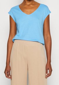 Světle modré tričko s krátkým rukávem a véčkovým výstřihem vyrobené z měkké látky, spojené s béžovými kalhotami s elastickým pasem. Jednoduchý design, uvolněný střih.