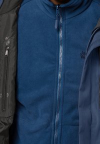 Niebieska polarowa kurtka z wysokim kołnierzem, pełnym zamkiem błyskawicznym i detalem z logo. Towarzyszy jej ciemnoszara warstwa zewnętrzna z akcentami zamka.