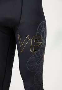 Schwarze Leggings mit einem großen, stilisierten "VE"-Logo in Orange und einem strukturierten Schlangendesign in Grau an der Seite. Glatte Stoffoberfläche.