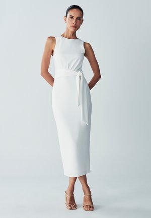 White sleeveless midi dress with a fitted bodice, tied waist detail, and smooth texture. Paired with nude block-heeled sandals.