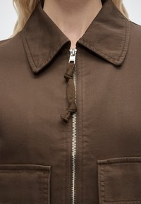 Καφέ ένδυμα με ασημένιο φερμουάρ στο μπροστινό μέρος και γιακά, που διαθέτει δύο εμπρόσθιες τσέπες και υφασμάτινη ταινία τράβηγματος φερμουάρ.
