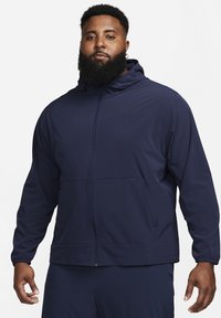 Marineblauwe Nike zip-up jas met capuchon, voorzien van een voorkanto en elastische manchetten, gemaakt van lichtgewicht, gladde stof.
