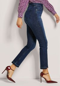 Donkerblauwe skinny jeans met een hoge taille, voorzien van een gladde textuur en subtiele stikseldetails; gecombineerd met bordeaux patent leren hakken.