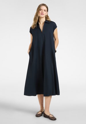 Frau trägt ein lockeres, dunkelblaues Midi-Kleid mit kurzen Ärmeln, Kragen, Seitentaschen und gewebten flachen Schuhen, steht vor einem schlichten Hintergrund.