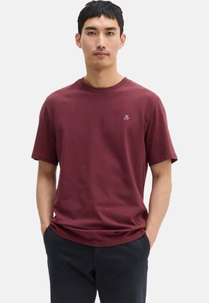 Mand med kort sort hår iført en bordeaux kortærmet t-shirt med lille hvidt logo og mørke bukser, står med hænderne i lommerne.