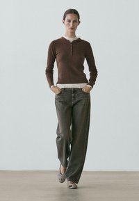 Young woman walking forward wearing brown long-sleeve shirt, gray wide-leg jeans, and beige patterned flats on a plain background.