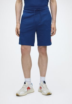 Homme portant un short bleu à cordon, des chaussettes blanches et des baskets blanches avec des accents rouges et noirs, debout devant un fond uni.