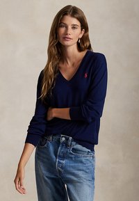 Ung kvinne med langt brunt hår iført en marineblå V-halsgenser med rød logo og lyseblå jeans, stående mot en ensfarget beige bakgrunn.