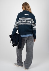 Personne portant un pull bleu marine à motifs, un jean gris et des baskets, tenant une veste sombre, dos à un fond uni.