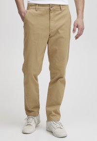 Chinos en coton beige avec une coupe droite, dotés d'une fermeture à bouton en métal et de poches latérales, associés à des baskets blanches.