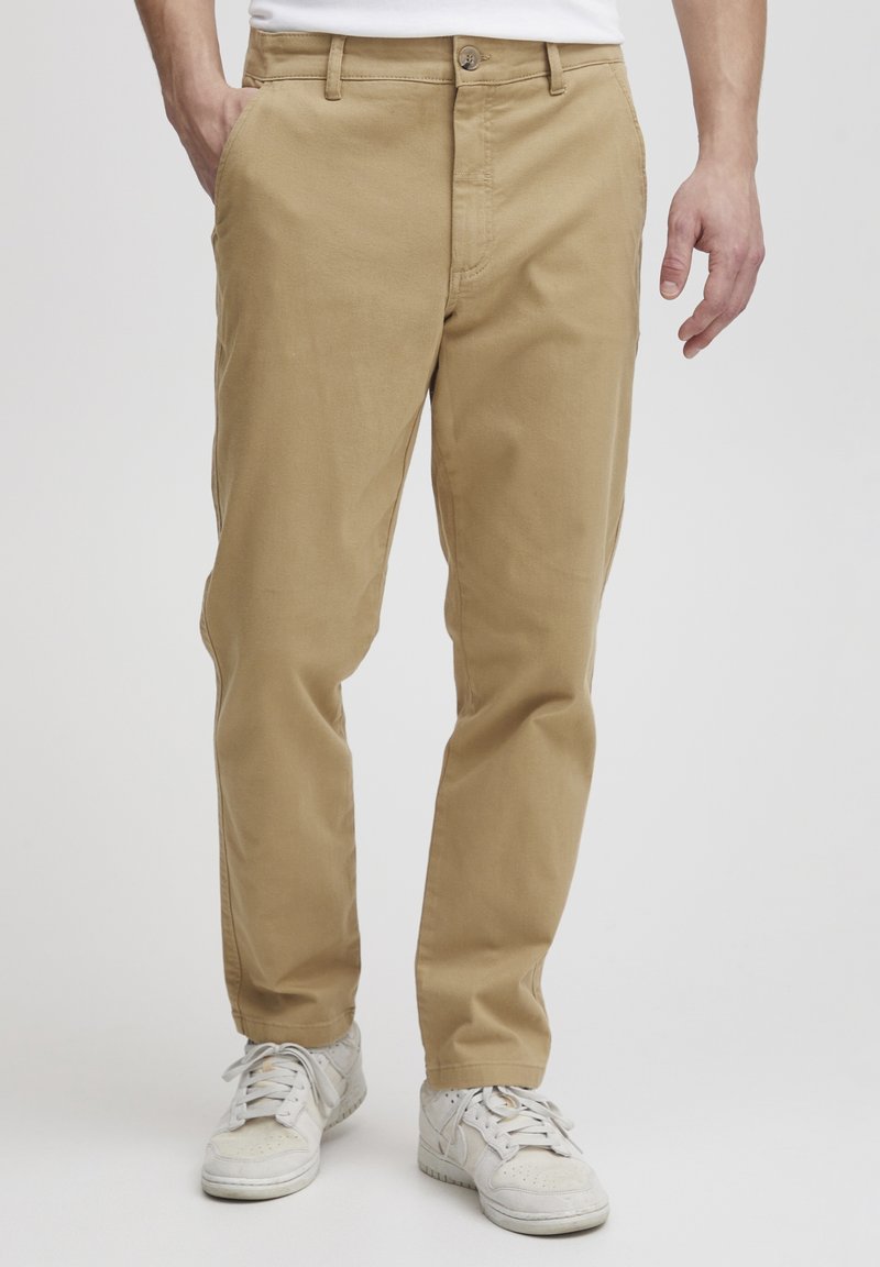 Chinos en coton beige avec une coupe droite, dotés d'une fermeture à bouton en métal et de poches latérales, associés à des baskets blanches.