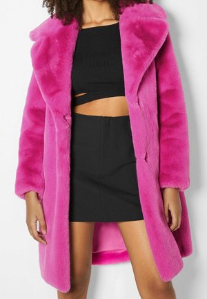 Femme portant un manteau en fausse fourrure rose vif par-dessus un crop top noir et une mini-jupe noire, debout devant un fond blanc.