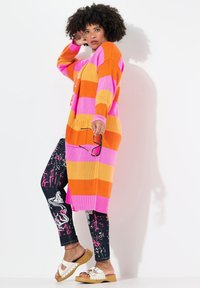Cardigan oversize a righe nei colori rosa, arancione e giallo. Leggings neri con motivo a macchie di vernice bianca. Sandali bianchi con piattaforma in legno.