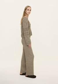 Beige kötött pulóver nyitott szövettel és hosszú ujjakkal, hozzáillő bőszárú nadrággal párosítva. A modellen fekete klumpák vannak. Simább semleges háttér.
