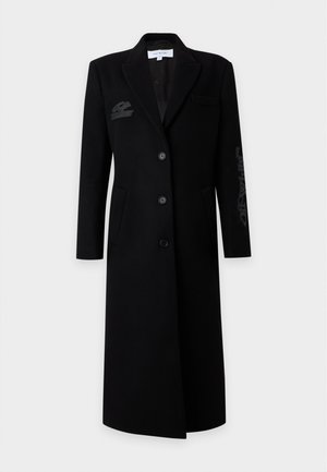 Femme aux longs cheveux portant un long manteau noir avec des boutons, debout les mains dans le dos sur un fond blanc uni.