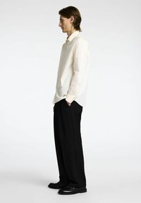 Camicia bianca con bottoni, pantaloni neri a gamba larga, scarpe nere lucide, tessuto morbido, vestibilità comoda, vista laterale del modello.