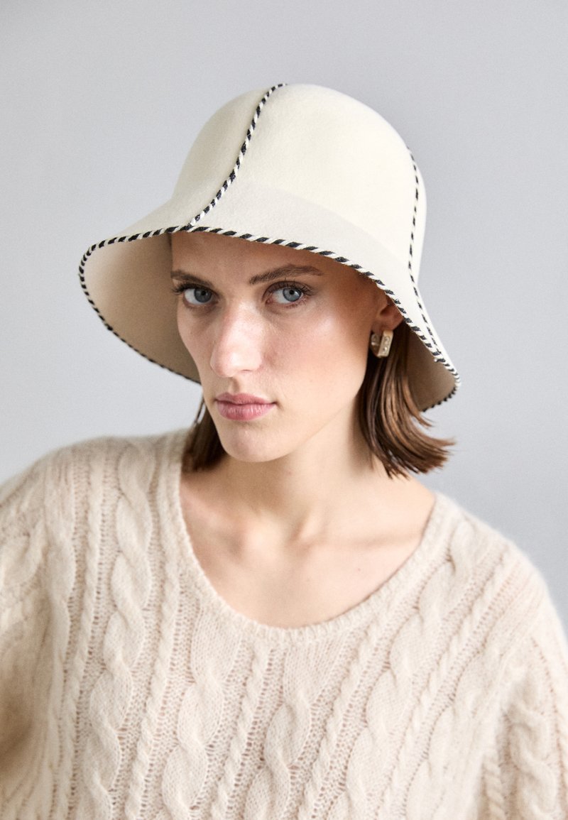 Chapeau en feutre beige avec un large bord, doté d'une couture noire contrastante sur les bords. Texture lisse et forme arrondie.