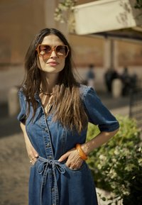 Ženska z dolgimi rjavimi lasmi, ki nosi velike kvadratne sončne naočnike in modro denim obleko, s rokama v žepih, stoji na prostem ob zelenju.