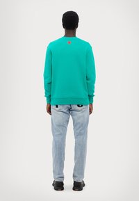 Felpa turchese con collo rotondo e logo rosso sul retro, abbinata a jeans blu chiaro e scarpe da ginnastica nere. Tessuto morbido.