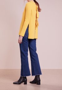Gelber gerippter Pullover mit langen Ärmeln und einem Rückenknotendesign. Kombiniert mit hochgeschnittenen, ausgestellten blauen Jeans und schwarzen, knöchelhohen Stiefeln mit Absatz.