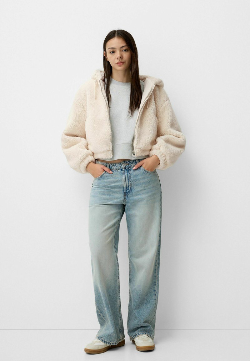 Veste en polaire crème coupée avec capuche, associée à un haut gris clair et un jean bleu taille haute. Des chaussures de sport complètent la tenue.