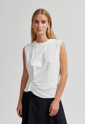 Femme aux longs cheveux blonds portant un haut blanc sans manches noué sur le côté et une jupe noire, debout devant un fond uni gris clair.