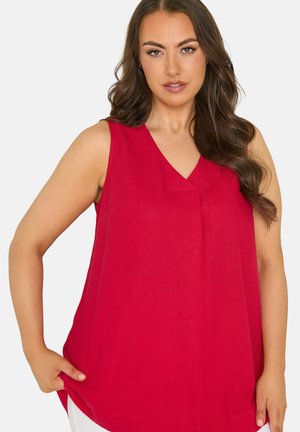 Kobieta o długich brązowych włosach, ubrana w czerwoną, bezrękawnikową bluzkę z dekoltem w serek, pozująca z jedną ręką trzymającą krawędź bluzki.