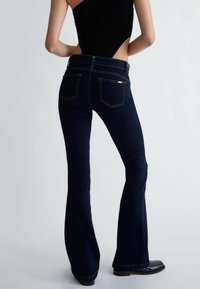 Los jeans de mezclilla oscuros de corte acampanado presentan una cintura alta, un diseño de cinco bolsillos, costuras visibles y un ligero brillo, combinados con una blusa negra de canalé.